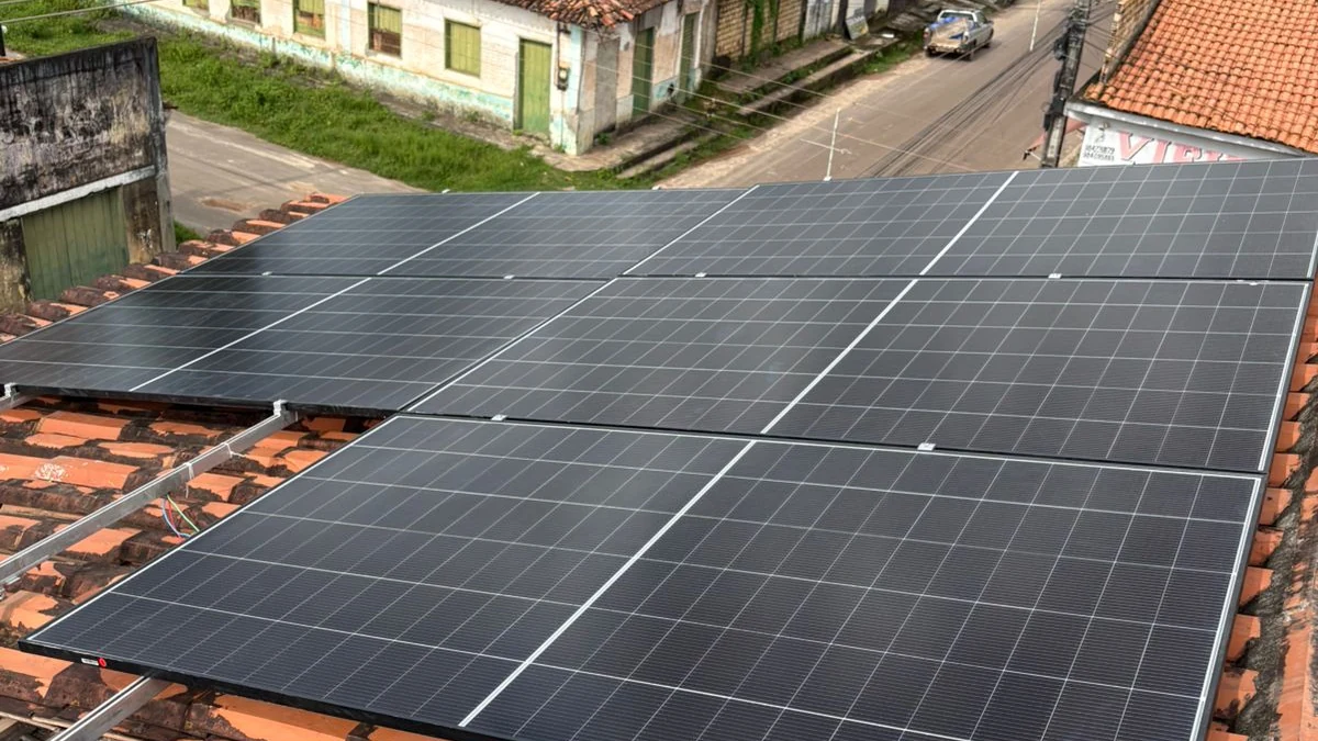 Painéis fotovoltaicos instalados sobre telhado cerâmico, vistos em plano amplo com parte da vizinhança ao fundo.